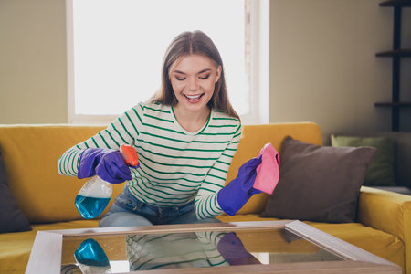 Young woman cleaning a glass table in a cozy living room under daylight, emphasizing a joyful and...