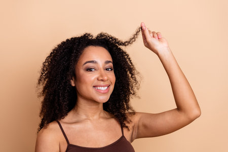 Young confident woman with curly hair smiling and touching her locks against a beige background celebrating casual beautyの写真素材