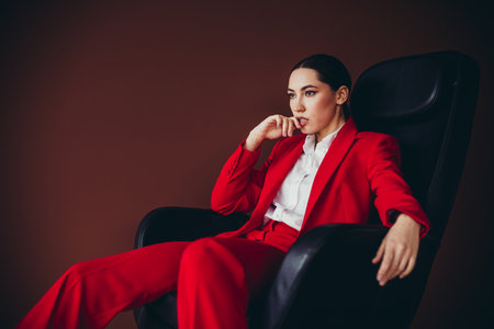 Beautiful Confident Businesswoman in Red Suit Sitting on Chair in Modern Office Settingの写真素材