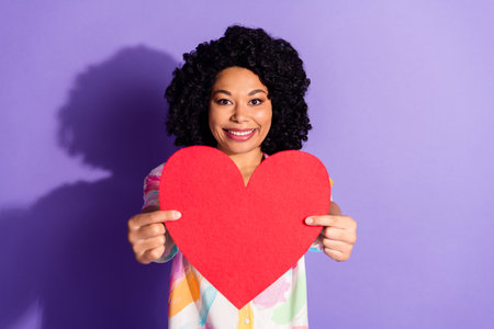 Young stylish woman holding large red heart symbol on purple background, representing love, happiness, and positive emotionsの写真素材
