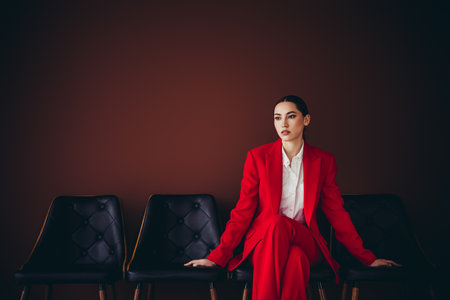 Stunning Confident Businesswoman Sitting Stylishly in Red Suit Against Brown Backgroundの写真素材