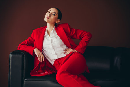 Stunning Elegant Businesswoman in Red Suit Relaxing on a Black Sofa in Modern Styleの写真素材