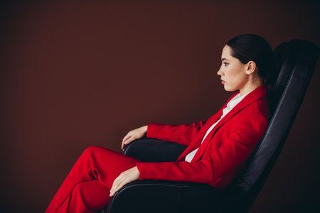 Stunning Elegant Businesswoman in Red Suit Sitting in Office Chair Against Brown Backgroundの写真素材