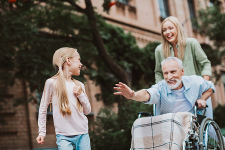 Funny cheerful moment as granddaughter greets grandpa in a wheelchair while mom smiles nearby in a sunny park sceneの写真素材