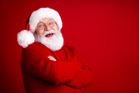 Joyful Santa Claus portrait in a warm red sweater and hat smiles with arms crossed against a bright red backgroundの写真素材
