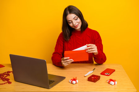 Cheerful young woman in red sweater opens a gift envelope beside a laptop on a bright yellow background enjoying holiday shopping momentの写真素材