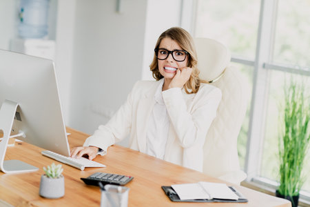 Worried businesswoman at desk using computer in modern office workspace with minimalistic design and green accentsの写真素材