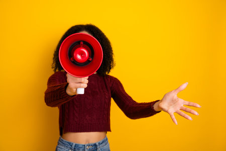 Young mixed race woman with curly hair holds a red megaphone and extends a hand against a bright yellow background promoting funの写真素材
