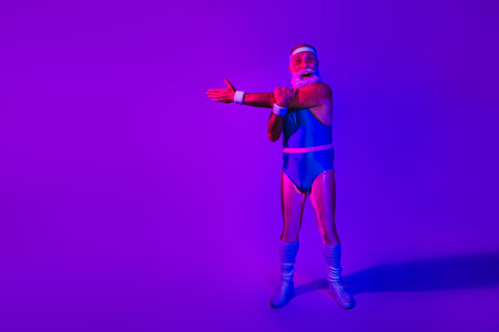 Elderly bearded man in retro blue jumpsuit poses with outstretched arms in neon purple and blue studio for fitness lifestyle and funの写真素材