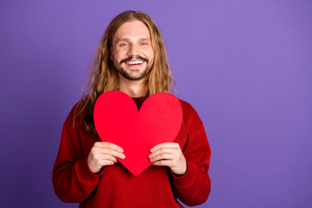 Young handsome man with long blonde hair smiles happily while holding a red heart against a purple background for love lifestyle and advertising visualsの写真素材