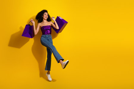 Young stylish woman jumping with purple shopping bags against yellow background capturing fashion energy joy and a playful shopping vibeの写真素材