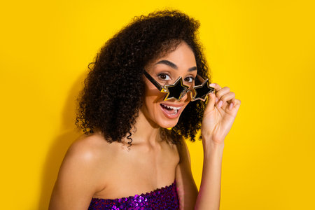 Young woman with star shaped sunglasses posing playfully against a bright yellow background for a...