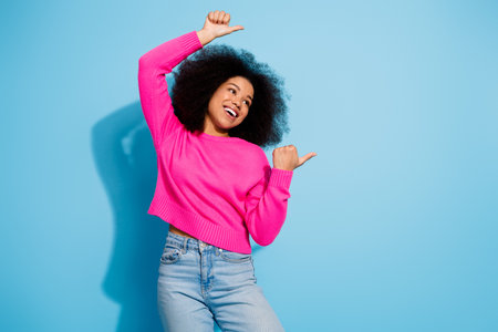 Young cheerful woman with black curly hair wearing a pink sweater on a light blue background showing...