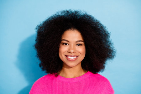 Portrait of a young woman in a vibrant pink sweater with an afro hairstyle and a cheerful expression on a blue backgroundの写真素材