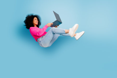 Young woman with curly hair using a laptop against a blue background, wearing a pink sweater and...
