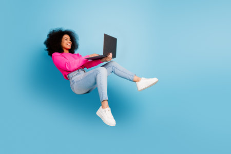 Cheerful young woman floating in air using laptop on vibrant blue background wearing pink sweater...