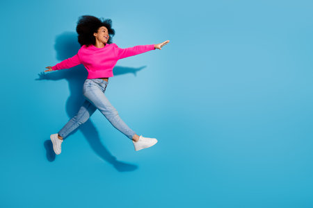 Cheerful young woman jumping against vibrant blue backdrop in stylish casual outfit showcasing...