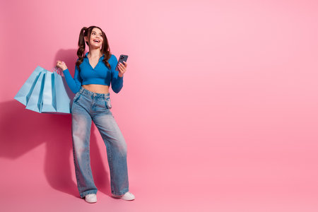 Cheerful young woman holding shopping bags and smartphone against a pink background, showcasing a vibrant and trendy lifestyleの写真素材