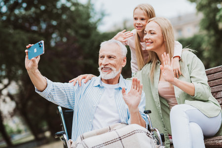 Joyful family moment as granddaughter and daughter take a selfie with grandpa in a wheelchair in the parkの写真素材