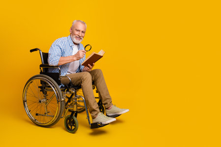 Grandpa in a wheelchair reads a book with a magnifying glass and smiles against a bright yellow background in a playful sceneの写真素材