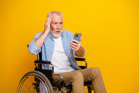 grandpa in wheelchair uses mobile phone with surprised expression yellow studio backdrop and casual checkered shirt conveys resilience and humorの写真素材