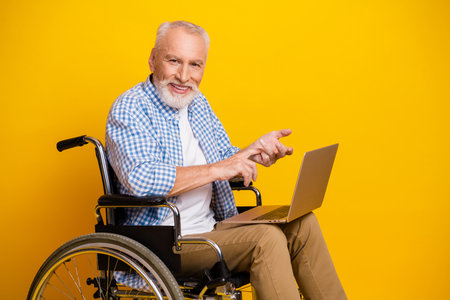 Cheerful grandpa in a wheelchair uses a laptop for learning and rehab with a bright yellow...