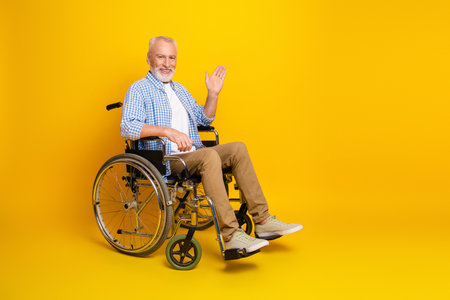 Happy senior grandpa in a wheelchair waving hello against a bright yellow background showcasing accessibility inclusionの写真素材