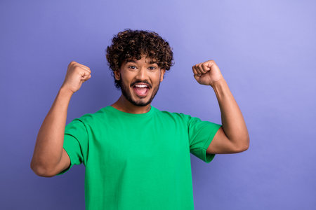 Cheerful young man in green t-shirt celebrating success with raised fists on vibrant purple backgroundの写真素材
