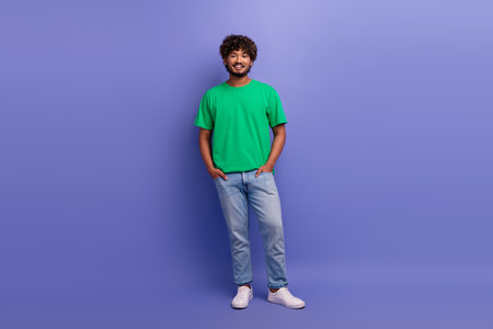 Young cheerful man in trendy green t-shirt and jeans standing against vibrant purple background, full body portraitの写真素材