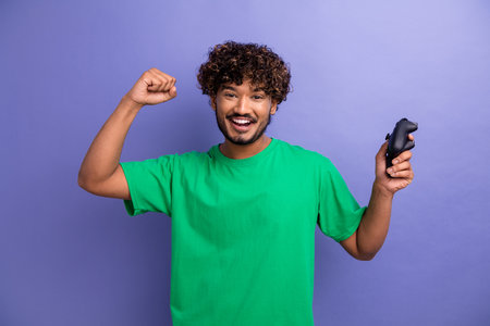 Young man enthusiastically celebrating while holding a game controller against a purple backdrop in casual green attireの写真素材