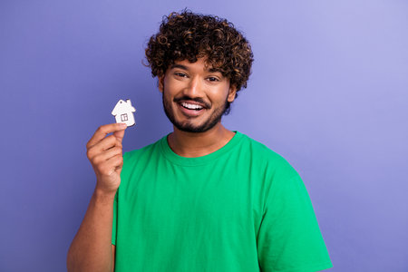 Cheerful young man holding a house-shaped item, wearing a green shirt against a vibrant violet backgroundの写真素材