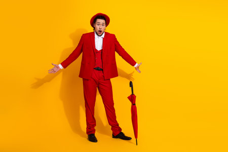 Funny stylish man in a red suit poses against a bright yellow background with an umbrella nearby for a bold fashion momentの写真素材
