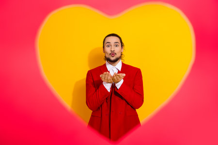 Funny man in a red suit blows a kiss in front of a giant yellow heart for a playful valentines day fashion momentの写真素材