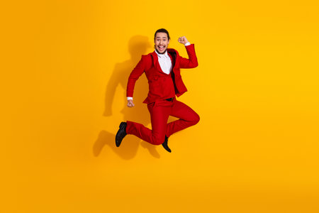 Energetic man in a red suit leaps across a bold bright yellow background creating a playful fashion momentの写真素材