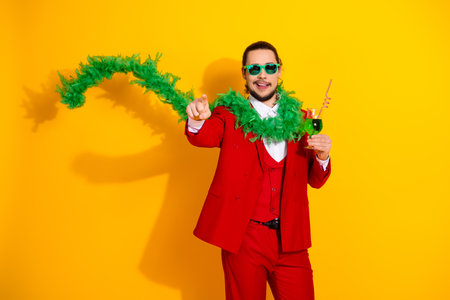 Vibrant man in red suit celebrates with green feather boa and drink against bright yellow background in a stylish festive momentの写真素材