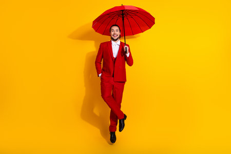 playful red suited model with umbrella leaps against yellow background for bold fashion and holiday style ad campaignの写真素材