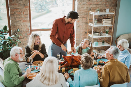 Fun and warm family gathering around the table as a cheerful host carves a turkey for grandparents parents and children during a joyful feastの写真素材
