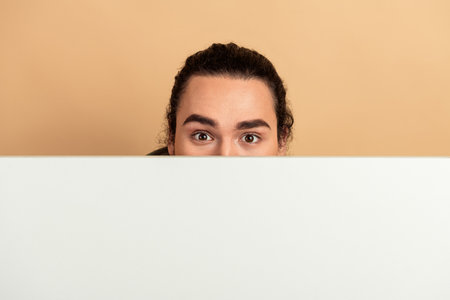Young man peeks over a white board with a beige background creating a playful casual lifestyle moment for fashion and advertising shootsの写真素材