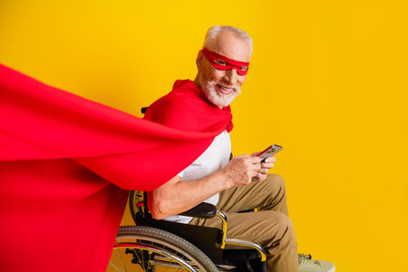 Playful senior superhero in wheelchair with red cape and phone strikes a joyful pose for a bright yellow studio sceneの写真素材