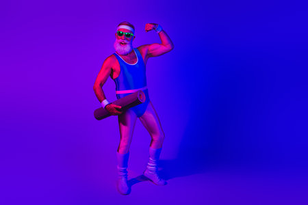 Retro fitness model with blue jumpsuit under neon purple lighting flexing arm and holding a mat in a studio portraitの写真素材