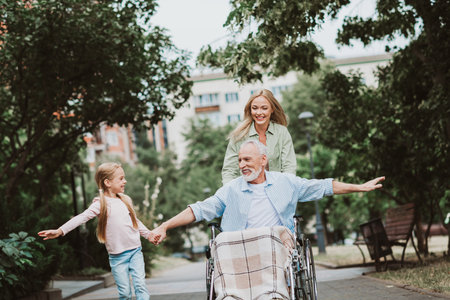 Joyful family moment as grandpa in wheelchair is cared for by daughter and wife in a sunny park walking together near trees and benchesの写真素材
