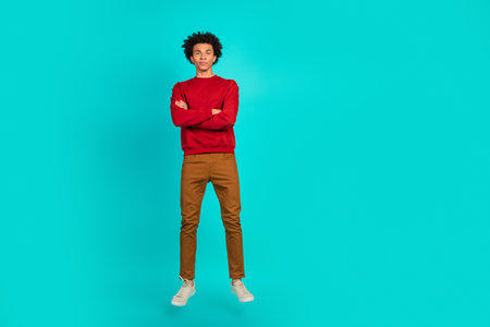 Young man in red sweater and brown pants stands with arms crossed against teal background showcasing casual fashion and modern style for lifestyle advertisingの写真素材