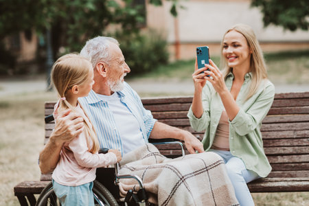 A heartwarming park selfie moment as grandpa in a wheelchair bonds with his granddaughter and loving daughter in sunshineの写真素材