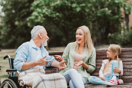 Heartwarming moment on a sunny park bench a grandpa in a wheelchair chats with his daughter and granddaughter sharing smiles and family careの写真素材