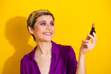 Young woman in purple blouse applying pink lipstick smiles at the camera against a bright yellow background for glamour fashion and beauty advertisingの写真素材