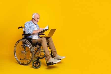 Grandpa in a wheelchair with headset and laptop smiles while writing and exploring online world against bright yellow background.の写真素材