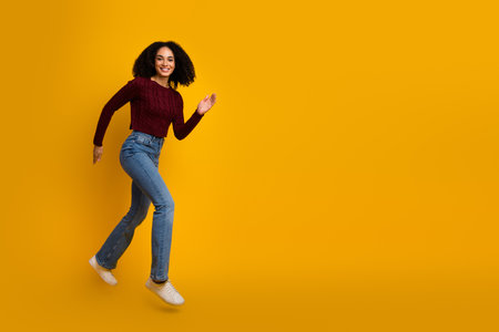 Young mixed race woman jumping against a vibrant yellow background wearing a burgundy sweater jeans and white sneakersの写真素材