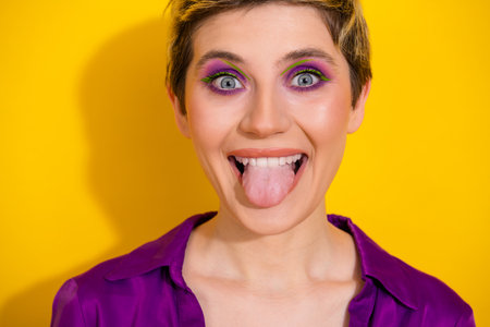Playful young woman sticks out tongue and smiles brightly against a bold yellow background wearing a purple shirtの写真素材