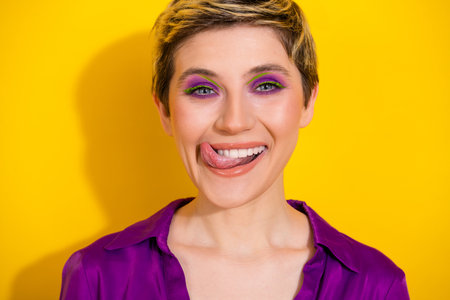 Playful young woman with bold makeup smiles and sticks out her tongue against a bright yellow background showcasing fashion beauty lifestyleの写真素材
