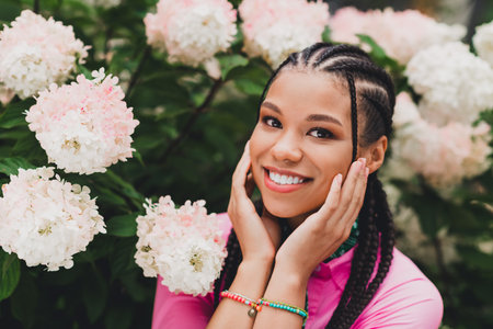 Young woman with braids smiles among pink and white hydrangeas in a garden wearing pink top and colorful braceletsの写真素材